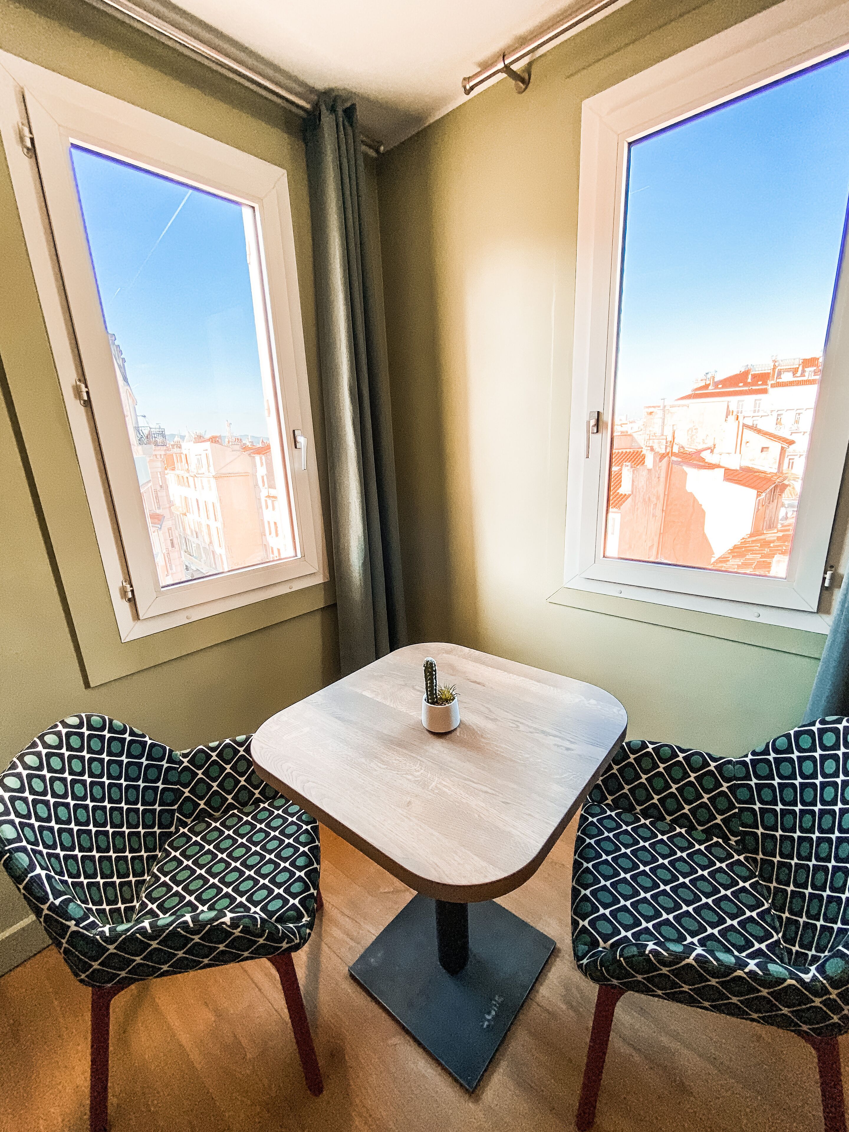 Une petite pièce avec une table et deux chaises à motifs, une fenêtre donnant sur des immeubles et un rideau.