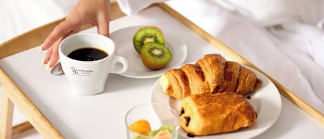 A tray with a croissant, kiwi slices, a cup of coffee, and a small glass with a dessert on a bed.