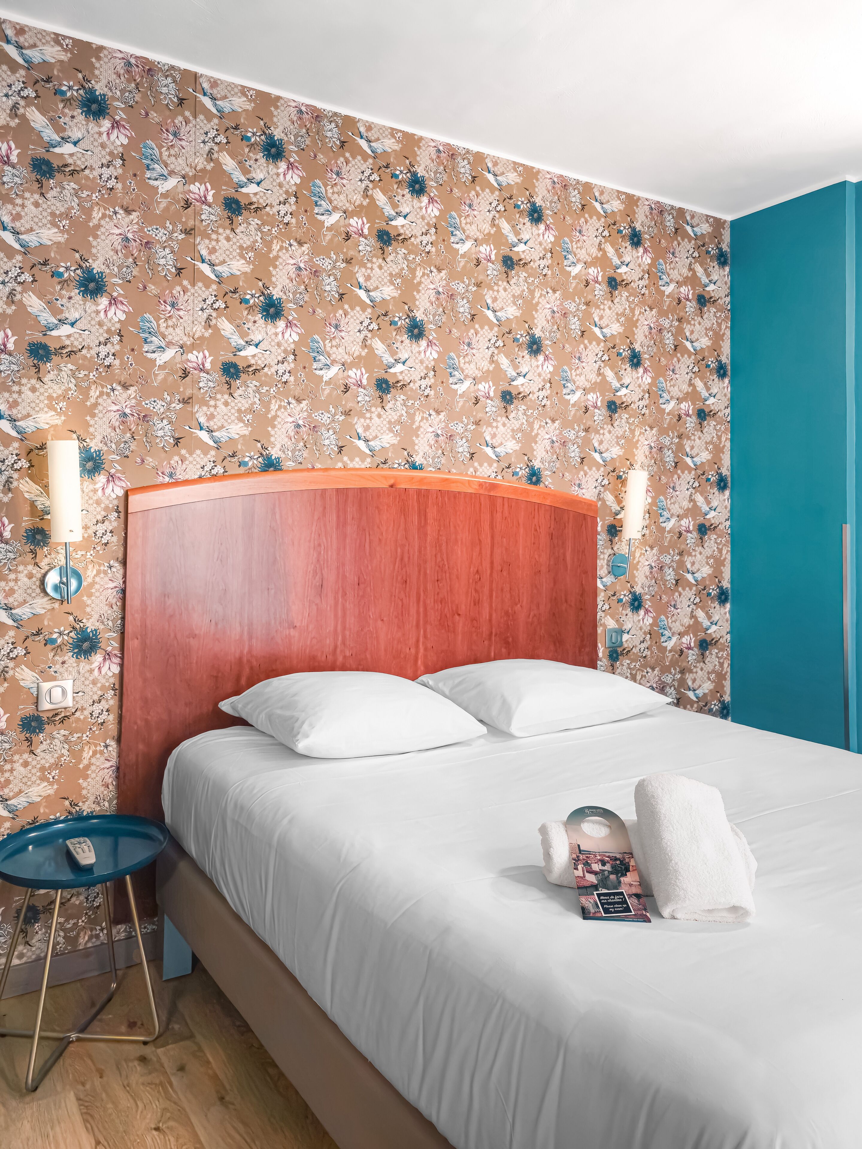 A hotel room with a floral wallpaper, a wooden headboard, white bedding, a bedside table with a lamp, and a blue stool.