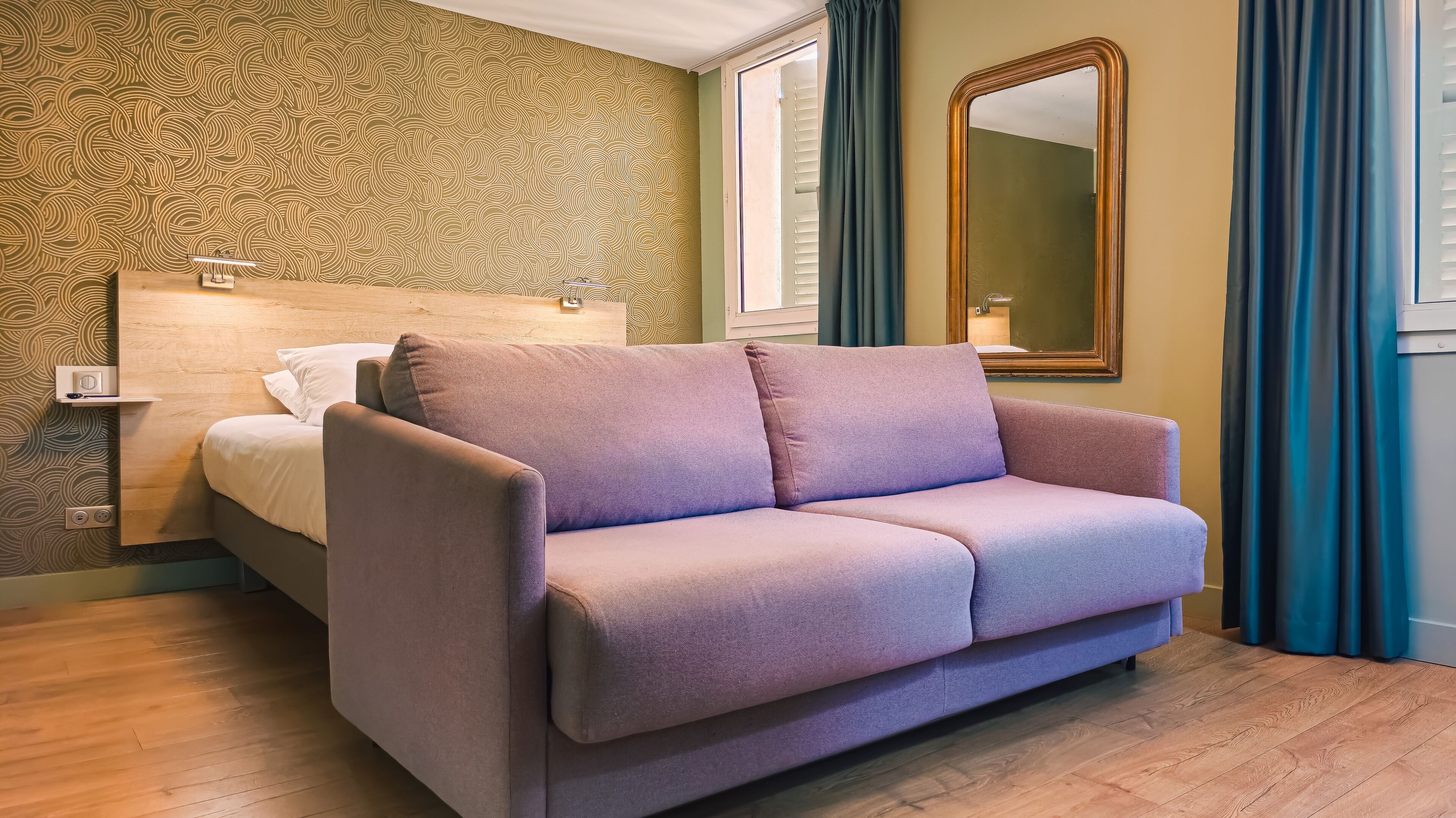 Una habitación con sofá morado, cama con cabecera de madera, un espejo grande y papel tapiz estampado.