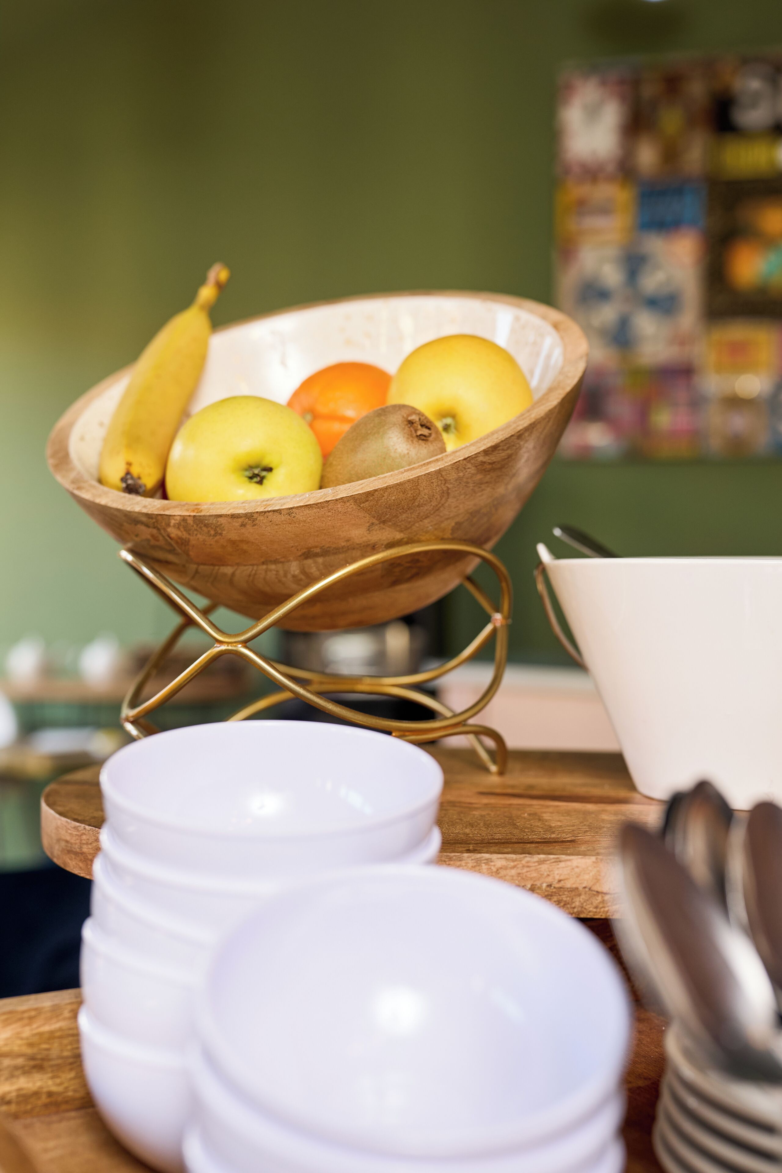 Un saladier en bois contenant des fruits, posé sur un présentoir, avec des assiettes blanches et des cuillères sur une table en bois.