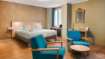 Habitación de hotel con una cama, dos sillones azules, una mesita redonda, un escritorio con una computadora y un espejo en la pared.