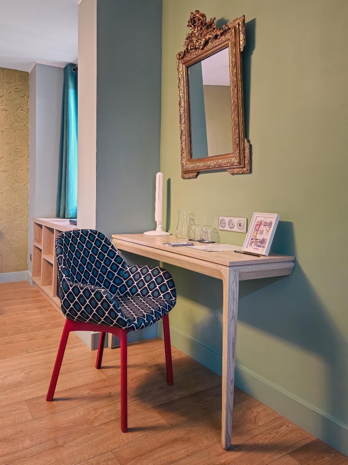 A room with a wooden desk, a chair, a mirror, and a wall with a patterned wallpaper.