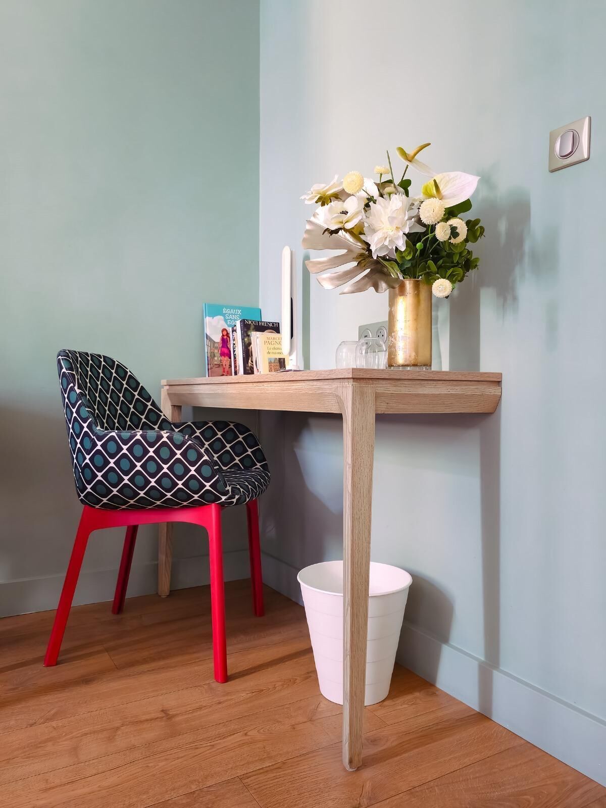 A corner room with a wooden desk, a patterned chair, a vase of flowers, and a white trash bin.
