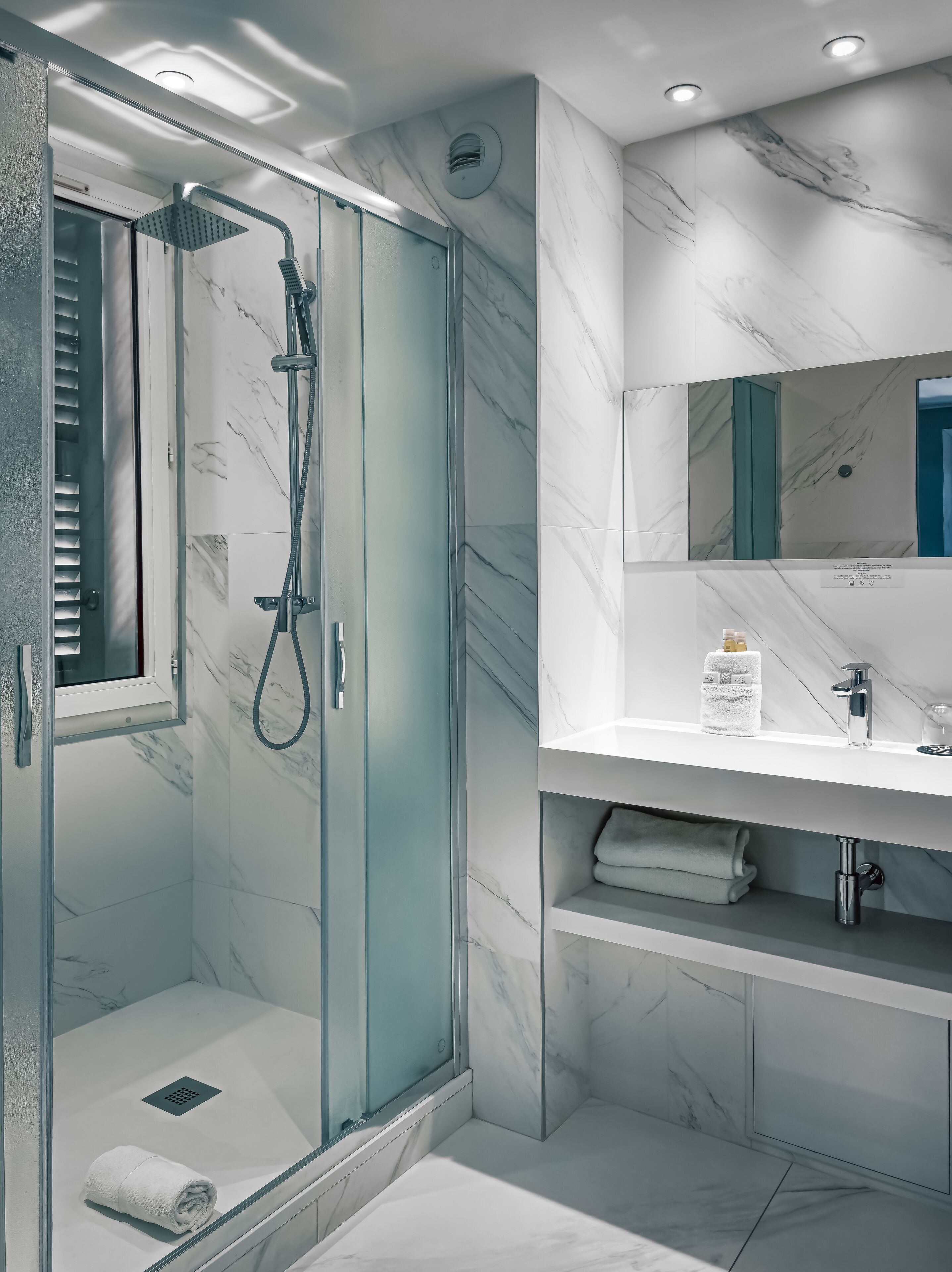 Une salle de bain moderne avec une douche à parois de verre, un mur recouvert de carreaux de marbre, un lavabo blanc et un miroir encastré.
