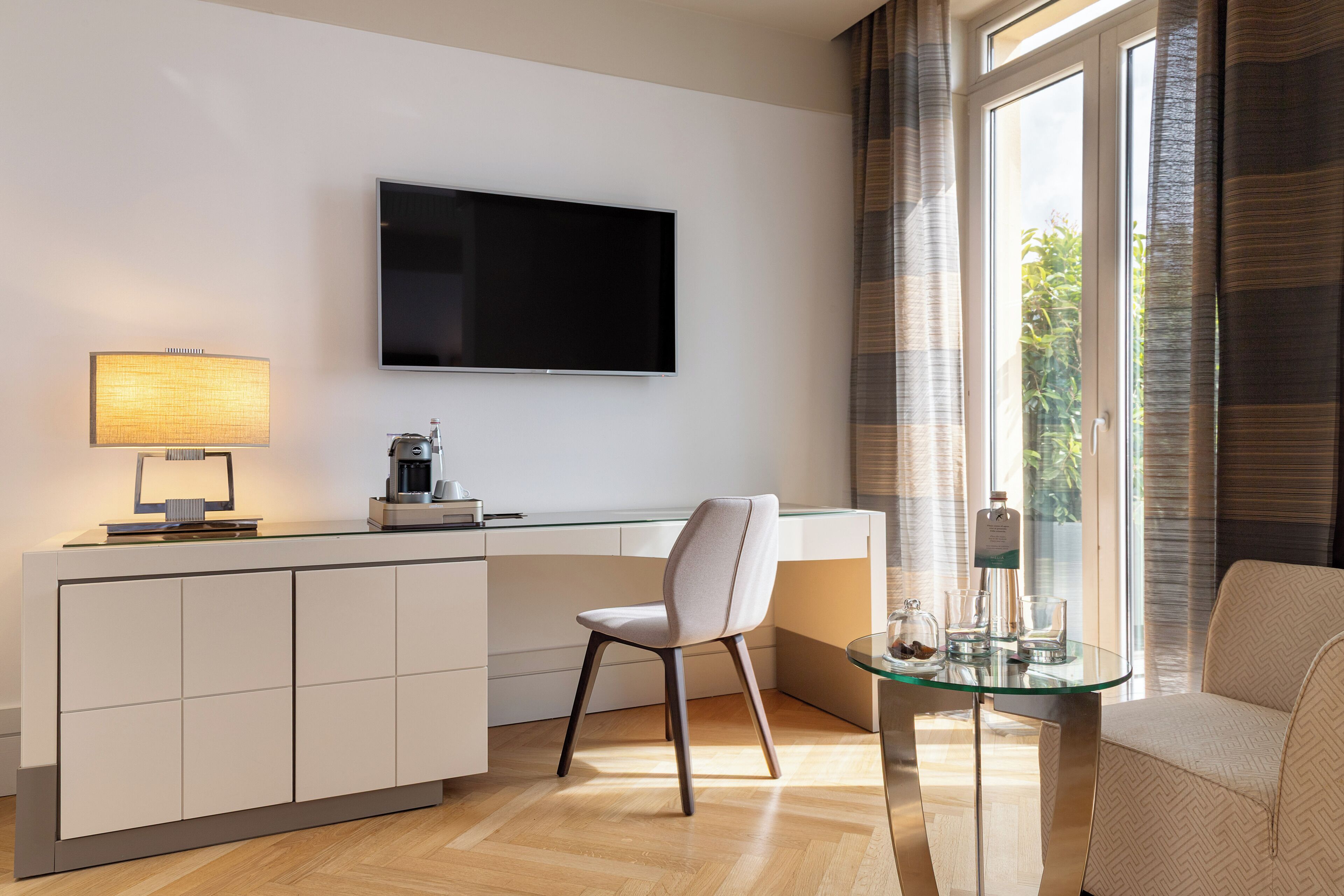 Une chambre d’hôtel moderne dotée d’un téléviseur à écran plat fixé au mur, d’une table à manger avec un plateau en verre, d’un canapé confortable et d’une grande fenêtre avec des rideaux.