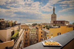 Una terraza con un plato de comida y una copa de vino, con vistas a un paisaje urbano que incluye edificios históricos y una torre de iglesia.