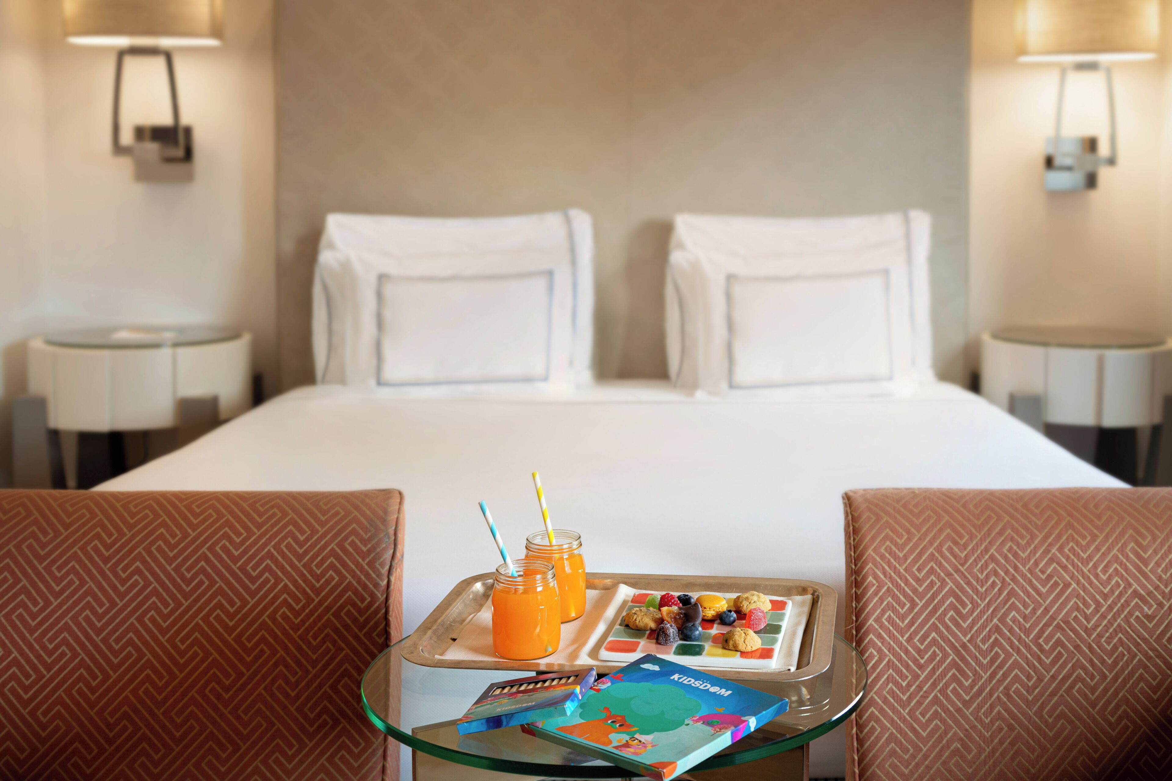 A hotel room with a neatly made bed, a breakfast tray with pastries and juice, and a children's book on a small table.