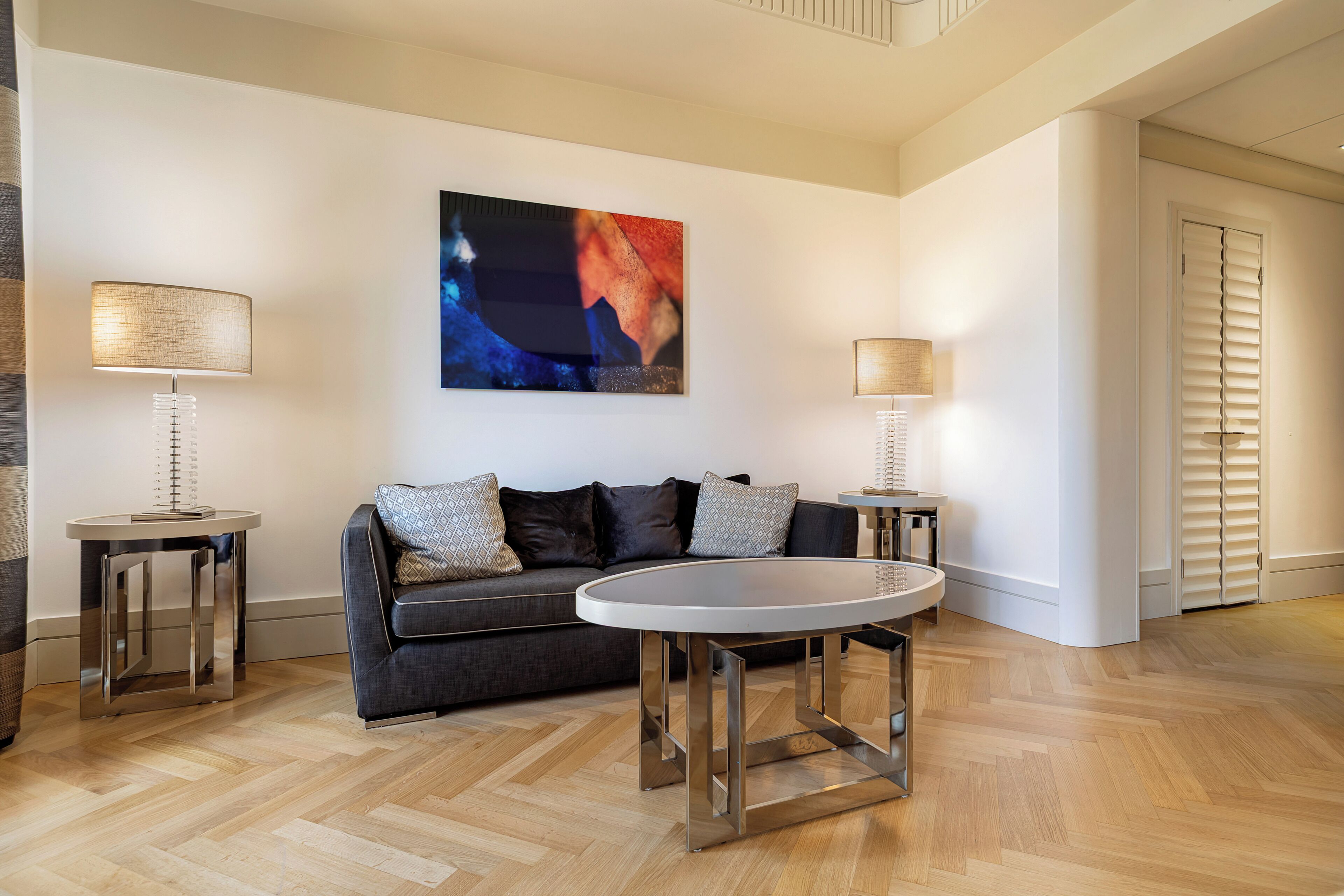 Una sala de estar moderna con un sofá gris oscuro, dos mesitas de noche con lámparas, una mesa redonda de centro y un cuadro abstracto grande en la pared.
