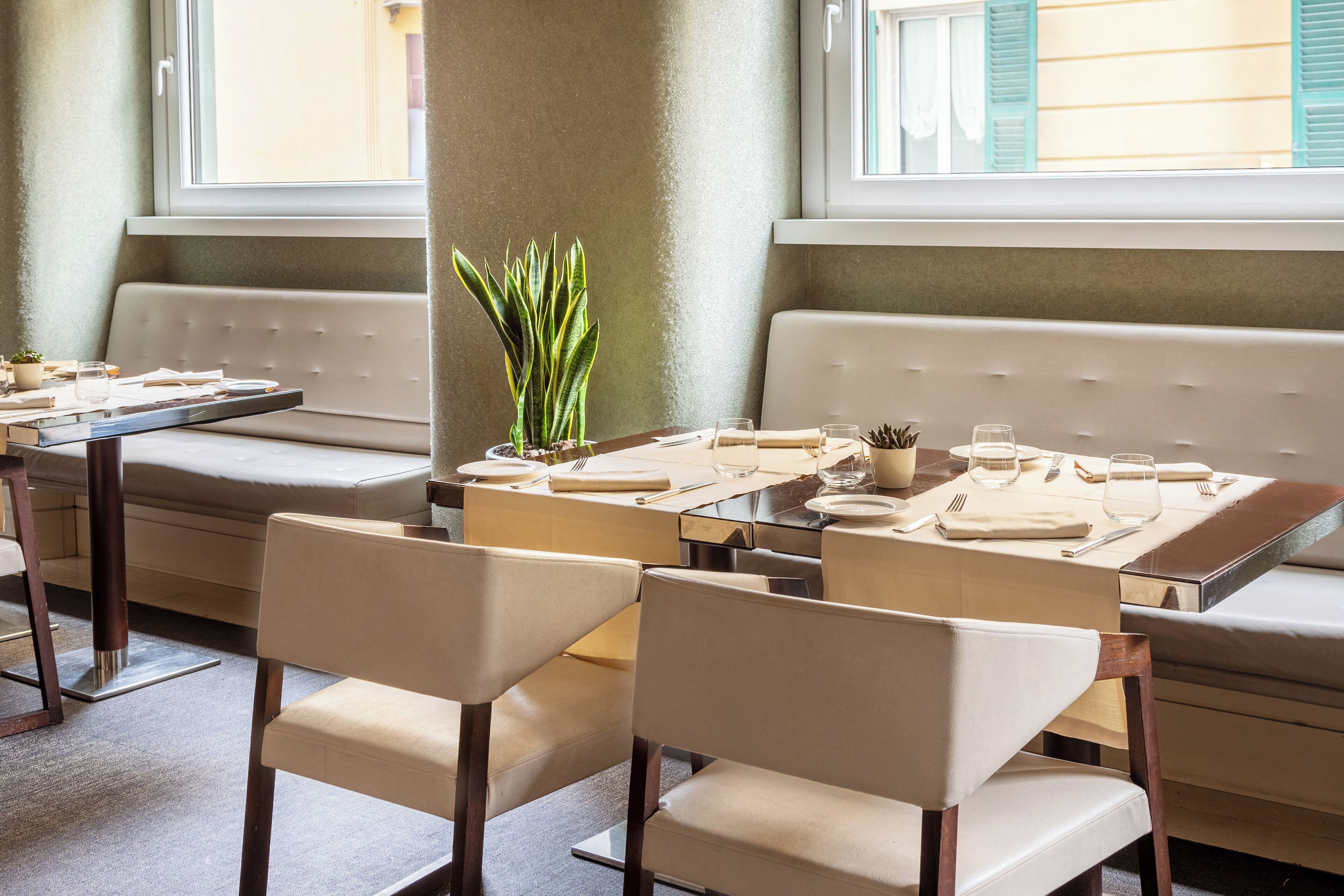 En restaurant med et langt bord dækket til spisning, polstrede siddepladser og en potteplante på bordet.