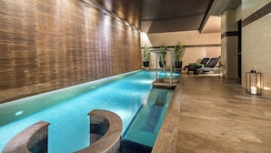 An indoor swimming pool with a tiled floor, a seating area with lounge chairs, and a wall with a textured pattern. - Melia Genova (Genoa)