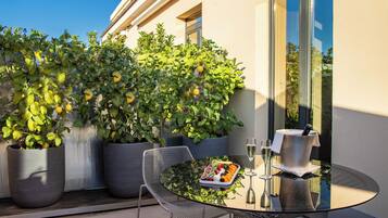 A glass table with a fruit tray and two glasses, surrounded by potted plants and a metal chair.
