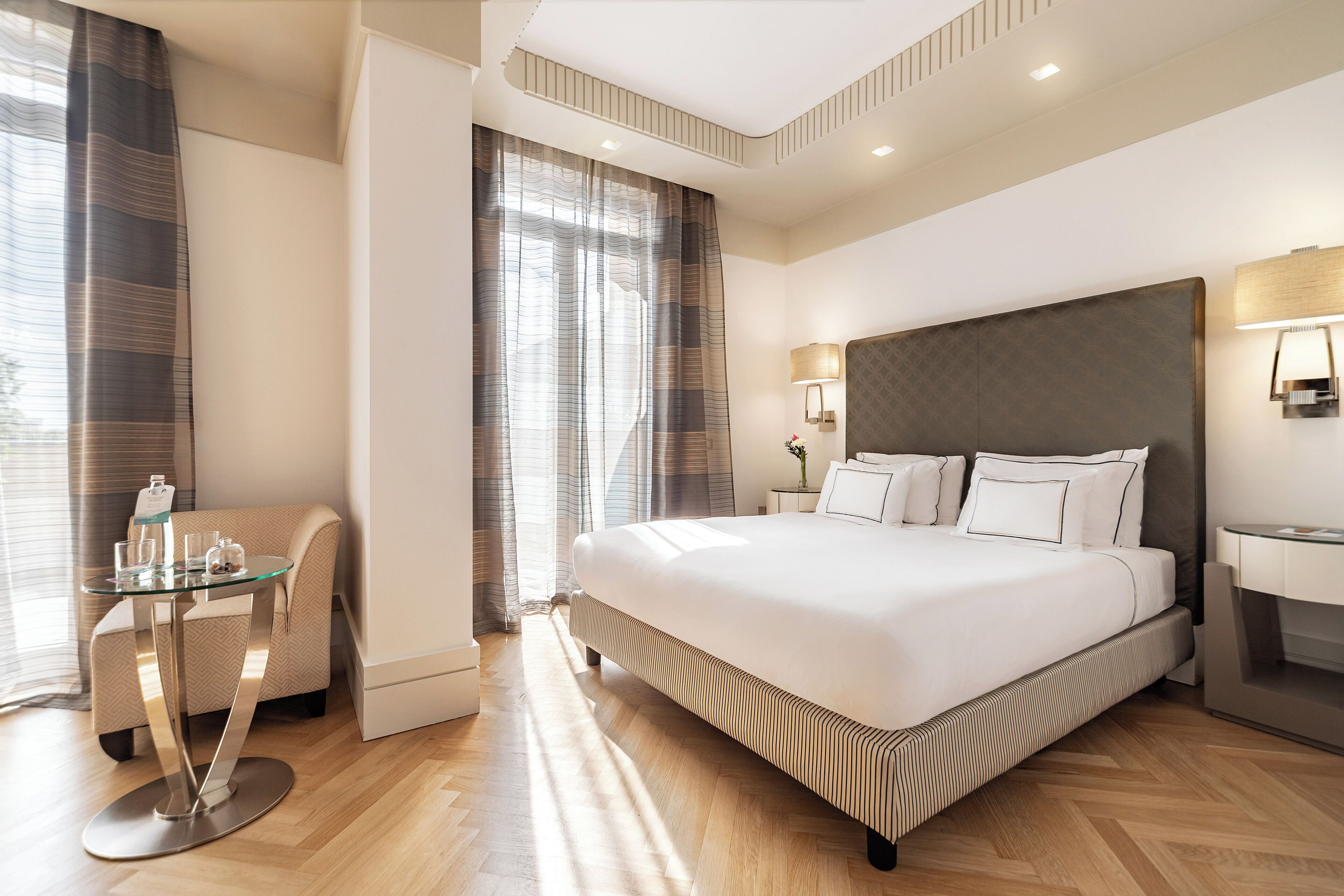 A modern hotel room with a large bed, a glass table, a chair, and a desk.