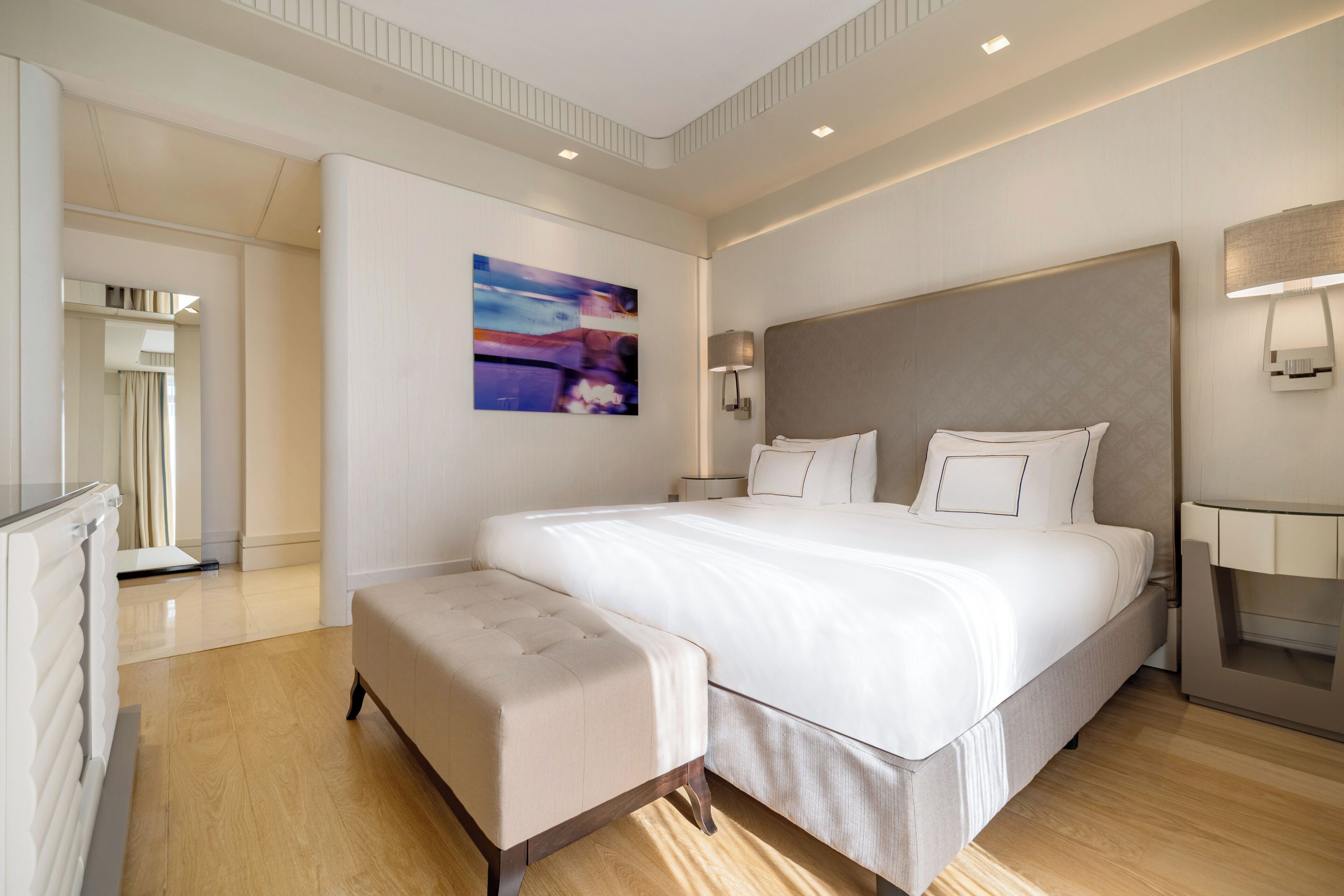 A modern hotel room with a large bed, a bedside table, a lamp, and a painting on the wall.