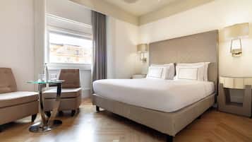 Habitación de hotel con una cama grande, una mesita de vidrio, dos sillones y vistas a edificios a través de una ventana.