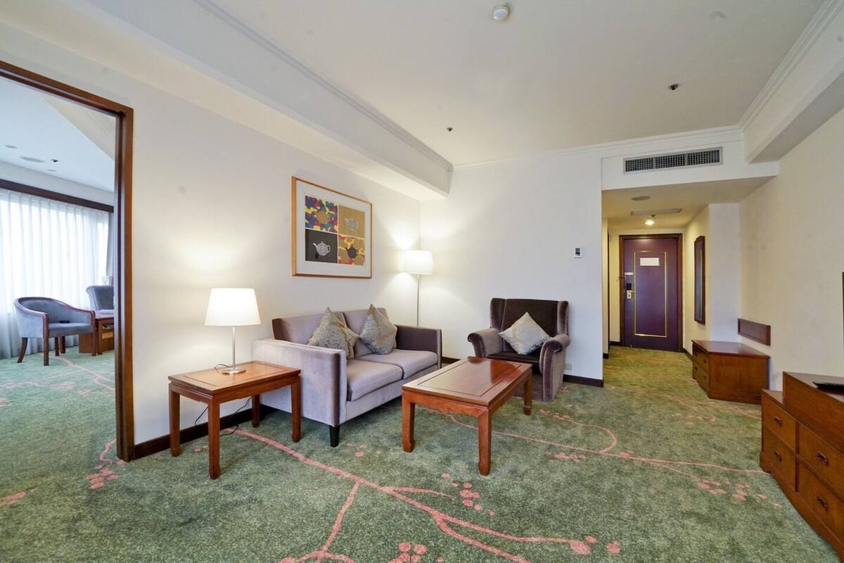 Una habitación de hotel con sofá, dos sillones, una mesa de centro, televisión y escritorio con lámpara.