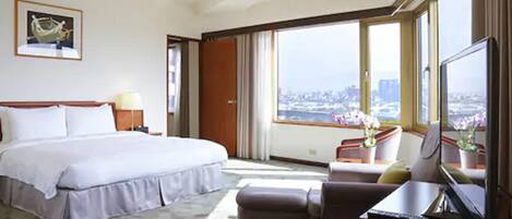 Habitación de hotel con una cama grande, un escritorio con computadora, una silla y vistas a un paisaje urbano a través de amplias ventanas.