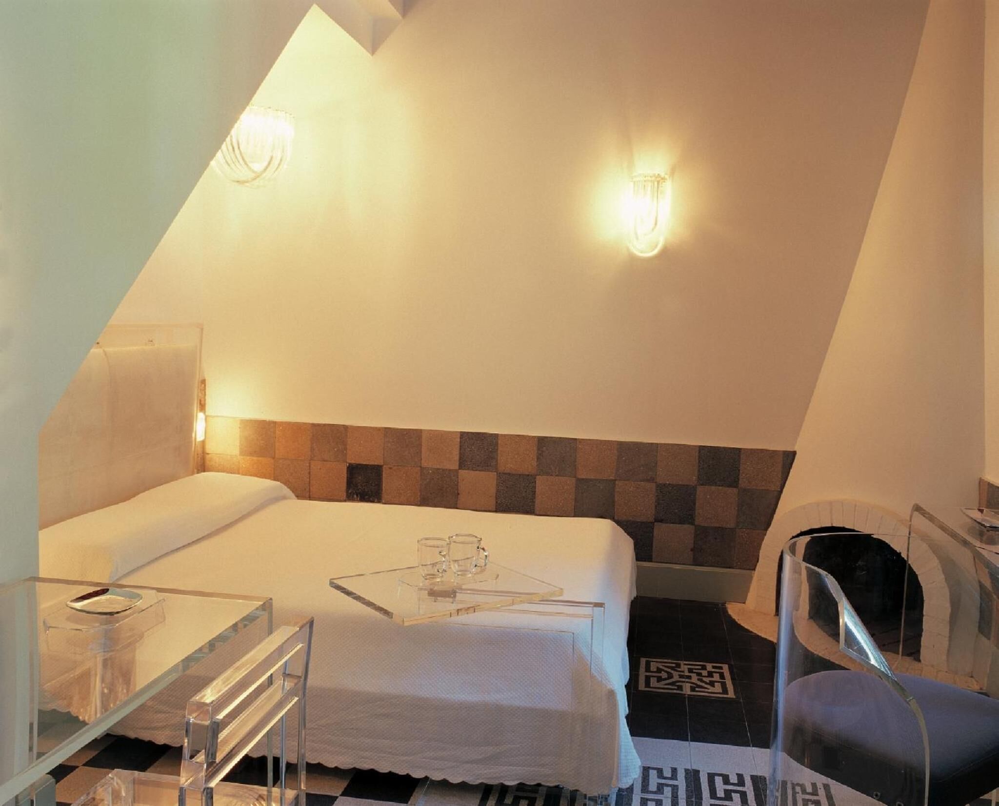Um quarto pequeno e triangular com uma cama, uma cadeira, uma mesa e duas luzes fixadas na parede.