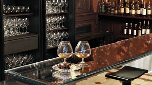 A bar area with shelves of wine glasses and bottles, two glasses of white wine on a glass table, and a chocolate dessert on a plate. - Villa Laetitia (Rome)