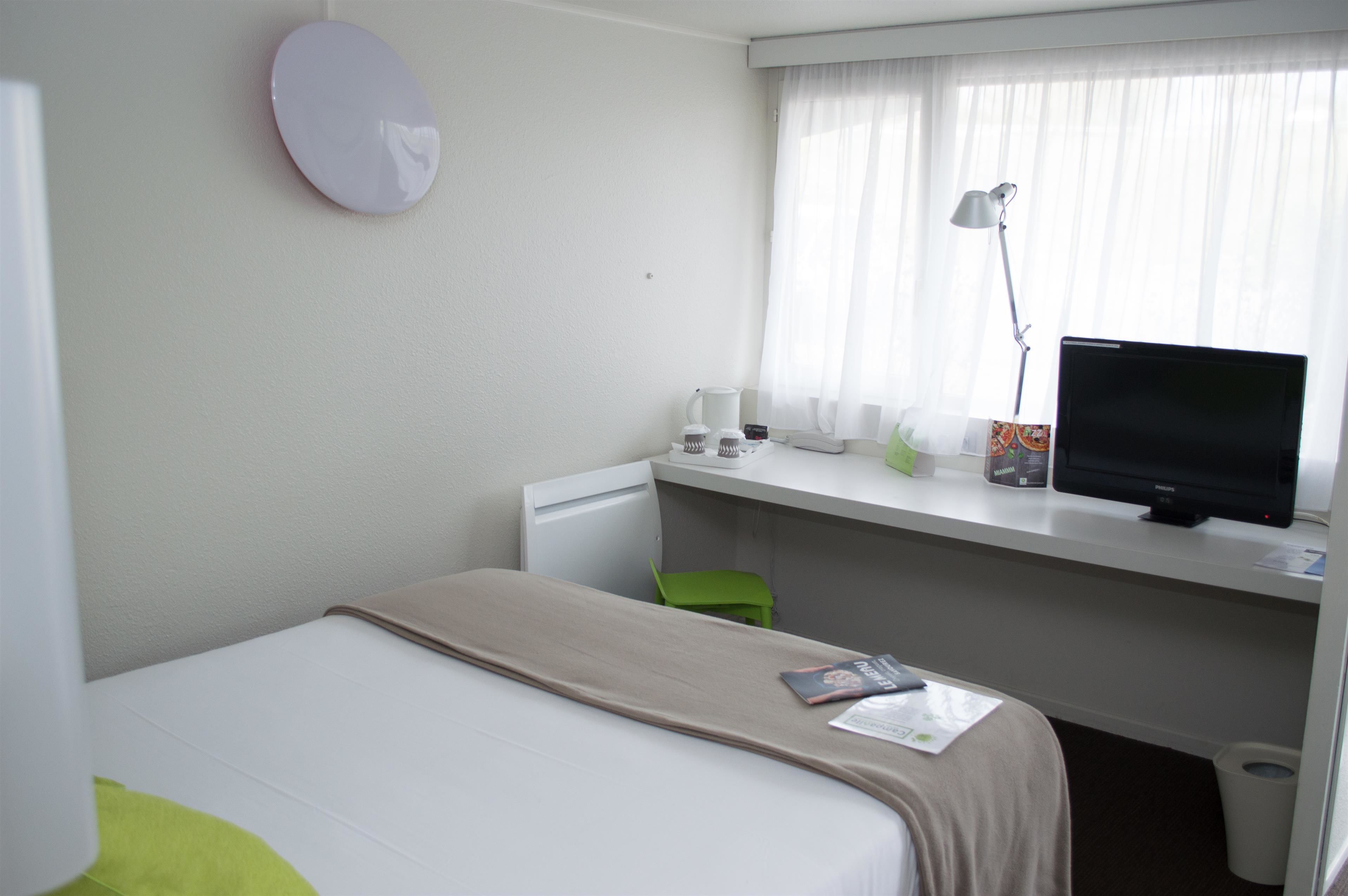 Quarto de hotel com uma cama, uma escrivaninha com computador, uma luminária e uma janela com cortinas.