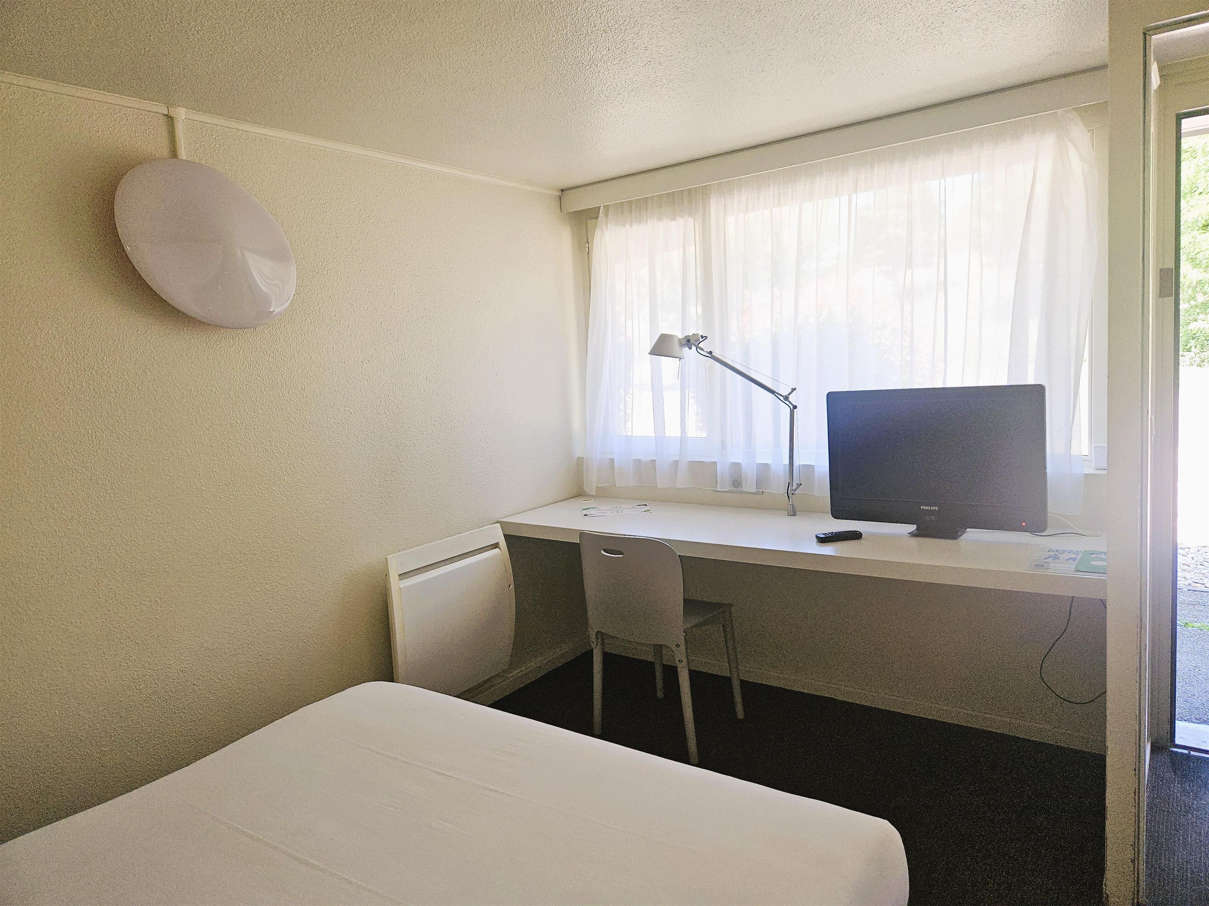 A small, clean room with a single bed, a desk with a computer and lamp, and a window with sheer curtains.
