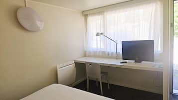 A small, clean room with a single bed, a desk with a computer and lamp, and a window with sheer curtains.