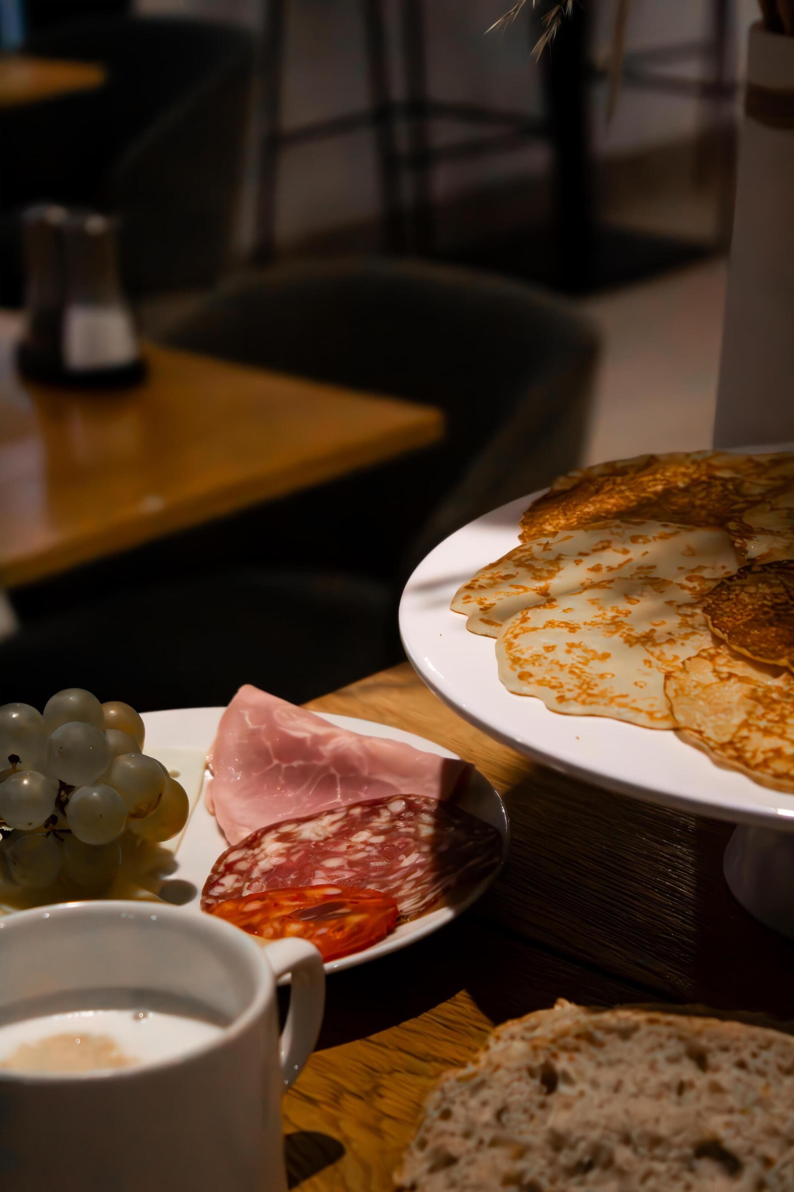 Prato com alimentos incluindo uvas, presunto e salame, uma xícara de café e uma fatia de pão sobre uma mesa de madeira.