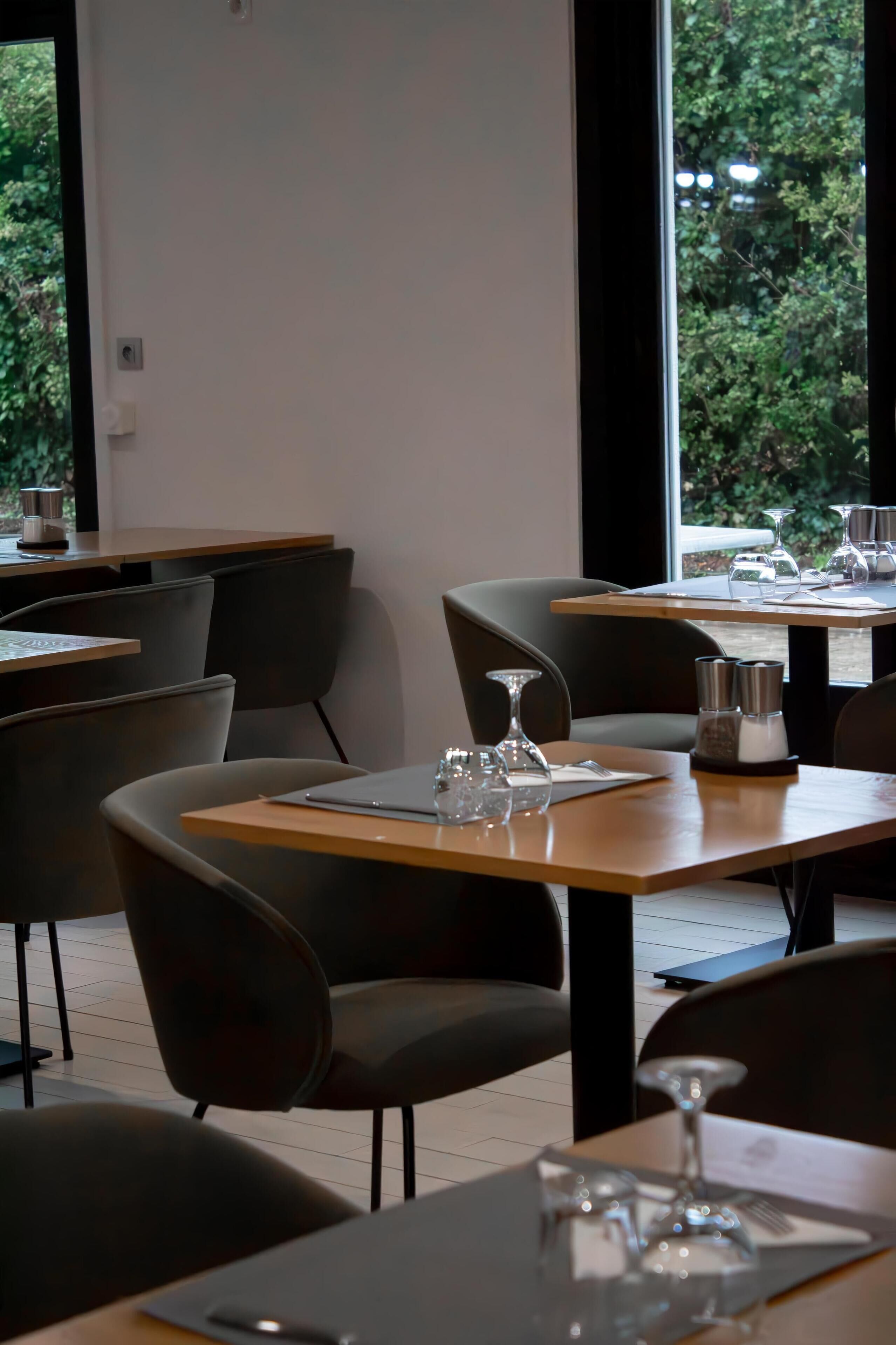 A modern dining area with tables set for two, featuring dark chairs and a view of greenery through large windows.