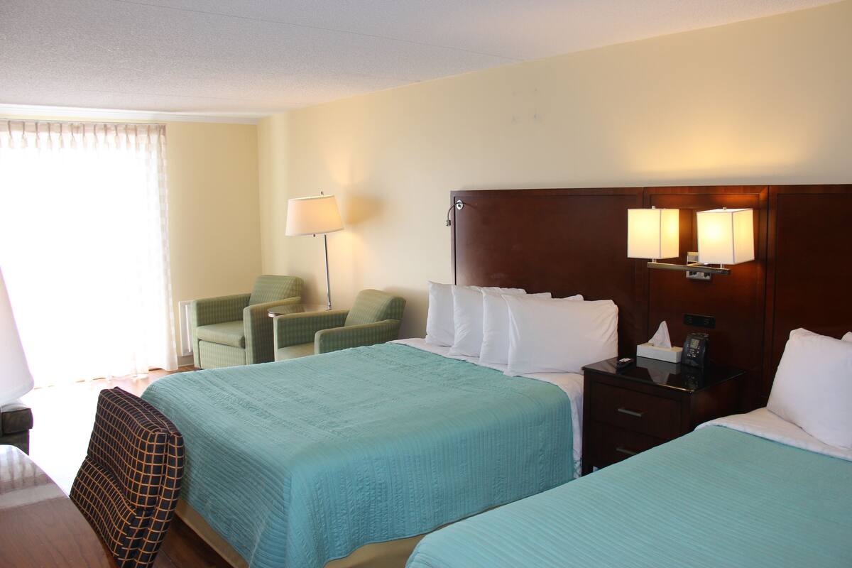 A hotel room with two beds, a desk, a chair, and a nightstand with a lamp.