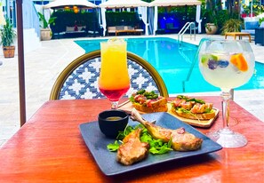 A dining table with a plate of grilled lamb chops, a glass of red wine, and a cocktail with fruit. In the background, there is a pool and outdoor seating. - Royal Palms Resort & Spa (Fort Lauderdale)