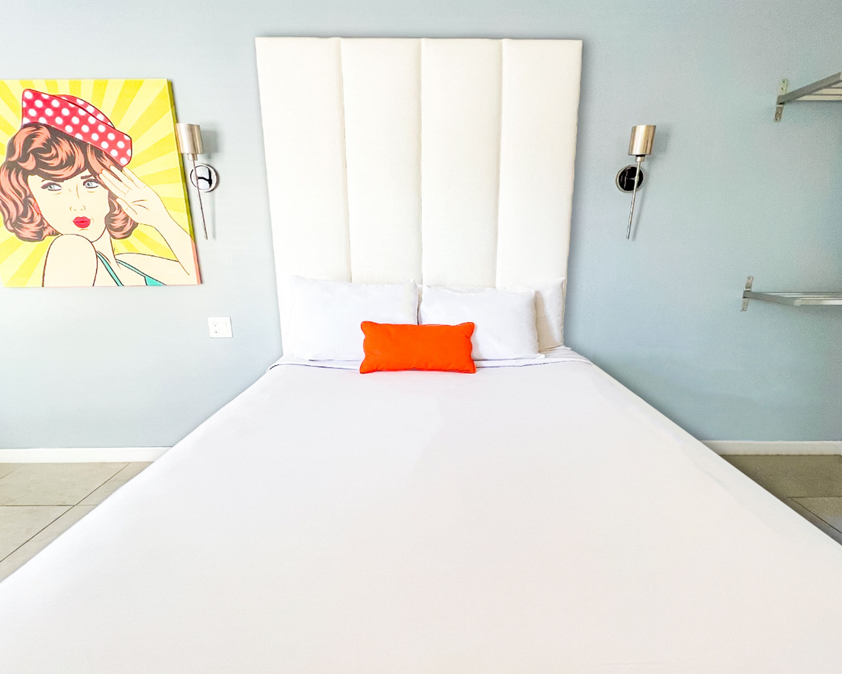 Ein Bett mit weißem Kopfteil und einem orangefarbenen Kissen, ein Gemälde einer Frau an der Wand und ein Nachttisch mit einer Lampe.