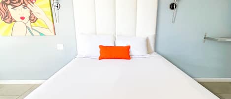 A bed with a white headboard and an orange pillow, a painting of a woman on the wall, and a bedside table with a lamp.