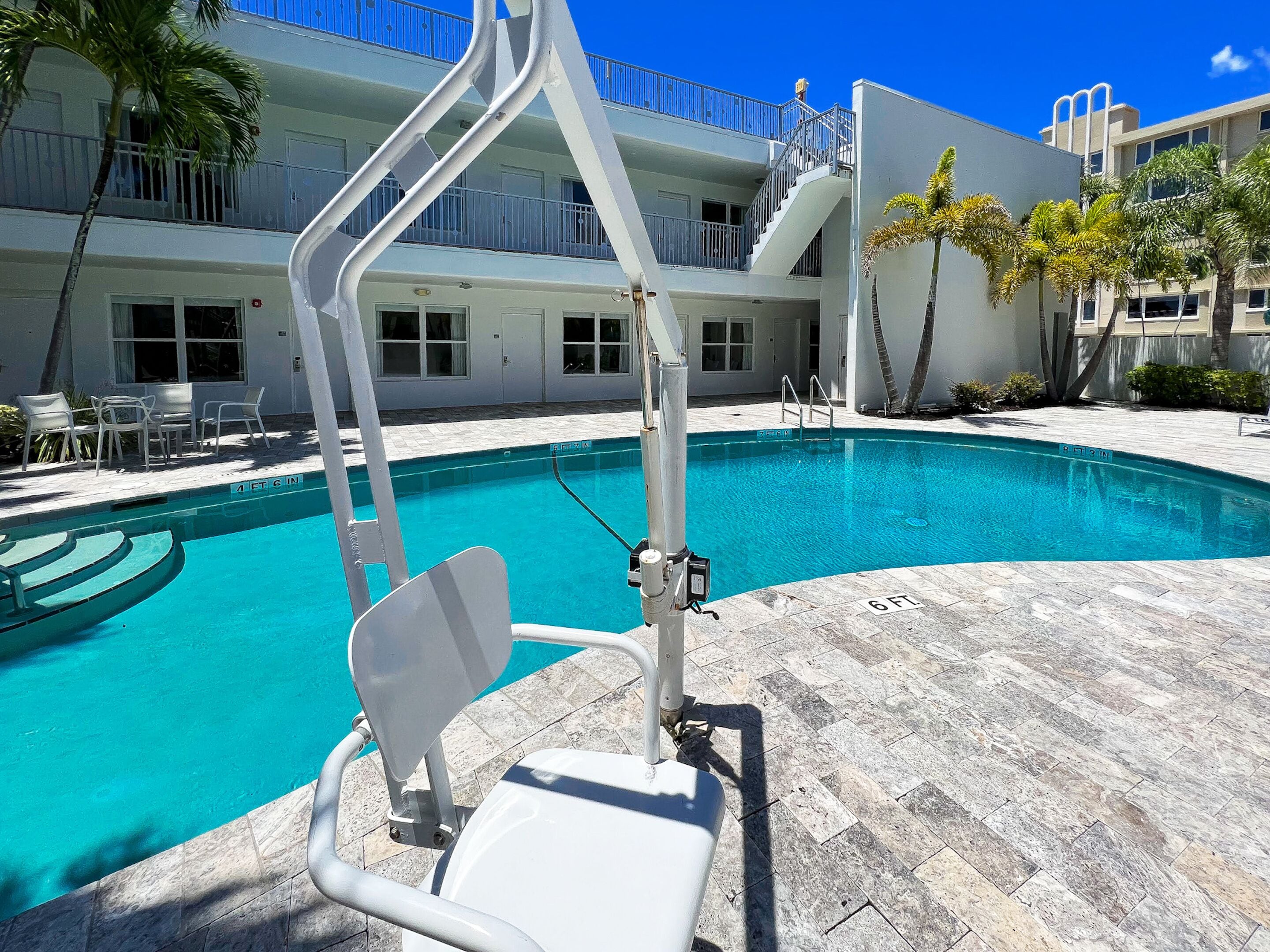 Une piscine avec un poste de surveillance pour le sauveteur, des chaises disposées au bord de la piscine et, au fond, un bâtiment doté de balcons.