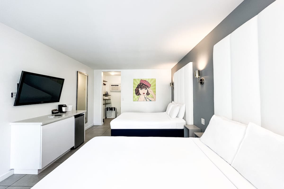A modern hotel room with a bed, a TV mounted on the wall, a small kitchenette, and a bathroom visible through an open door.