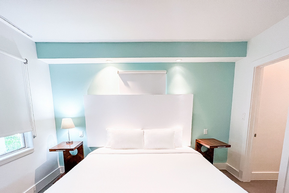 A hotel room with a large bed, a nightstand, a bedside table, and a window with blinds.