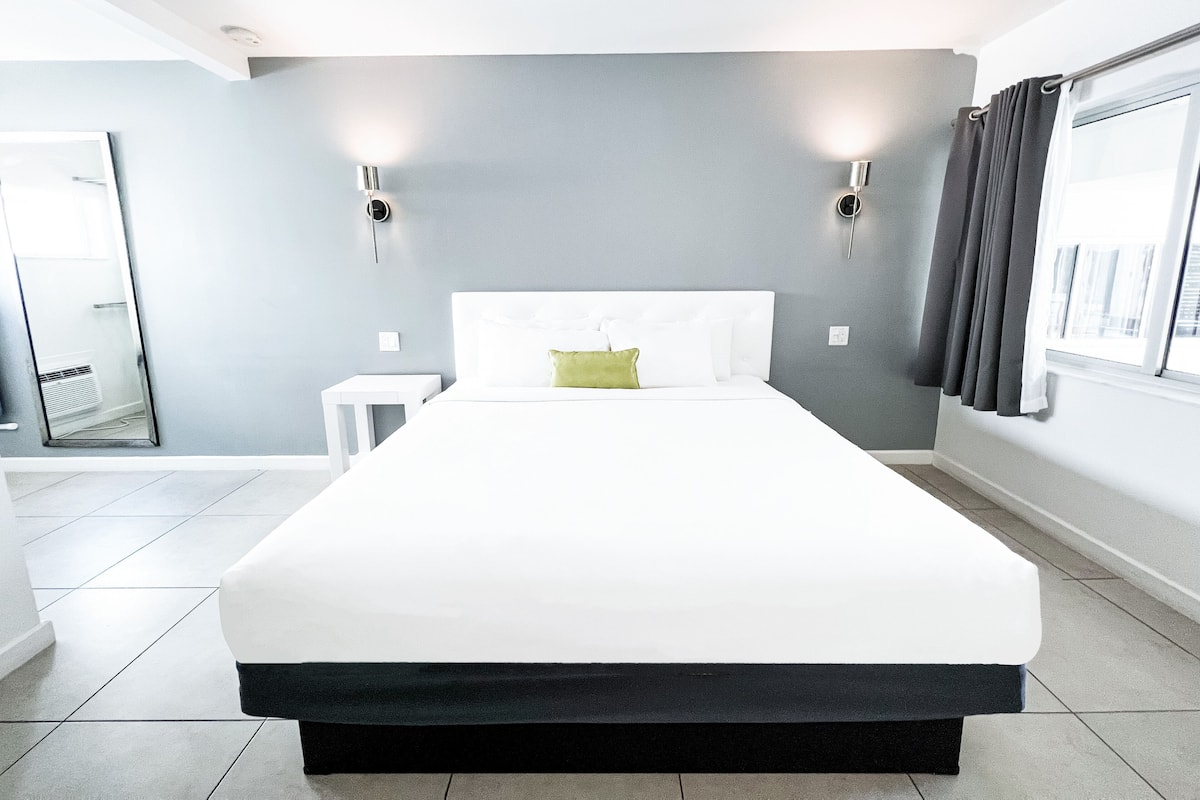 A modern hotel room with a large bed, a small bedside table, and a window with curtains.