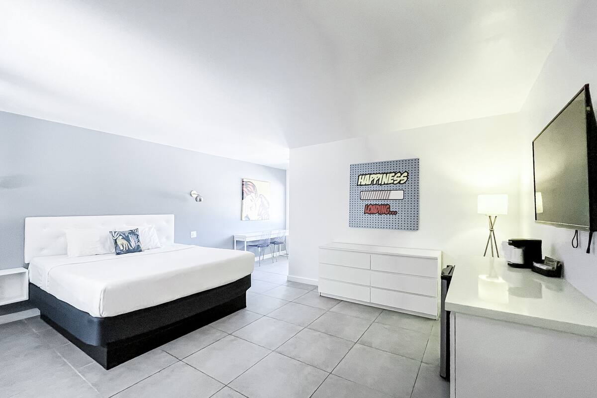 A modern hotel room with a large bed, a desk, a chair, a TV, and a wall art piece.