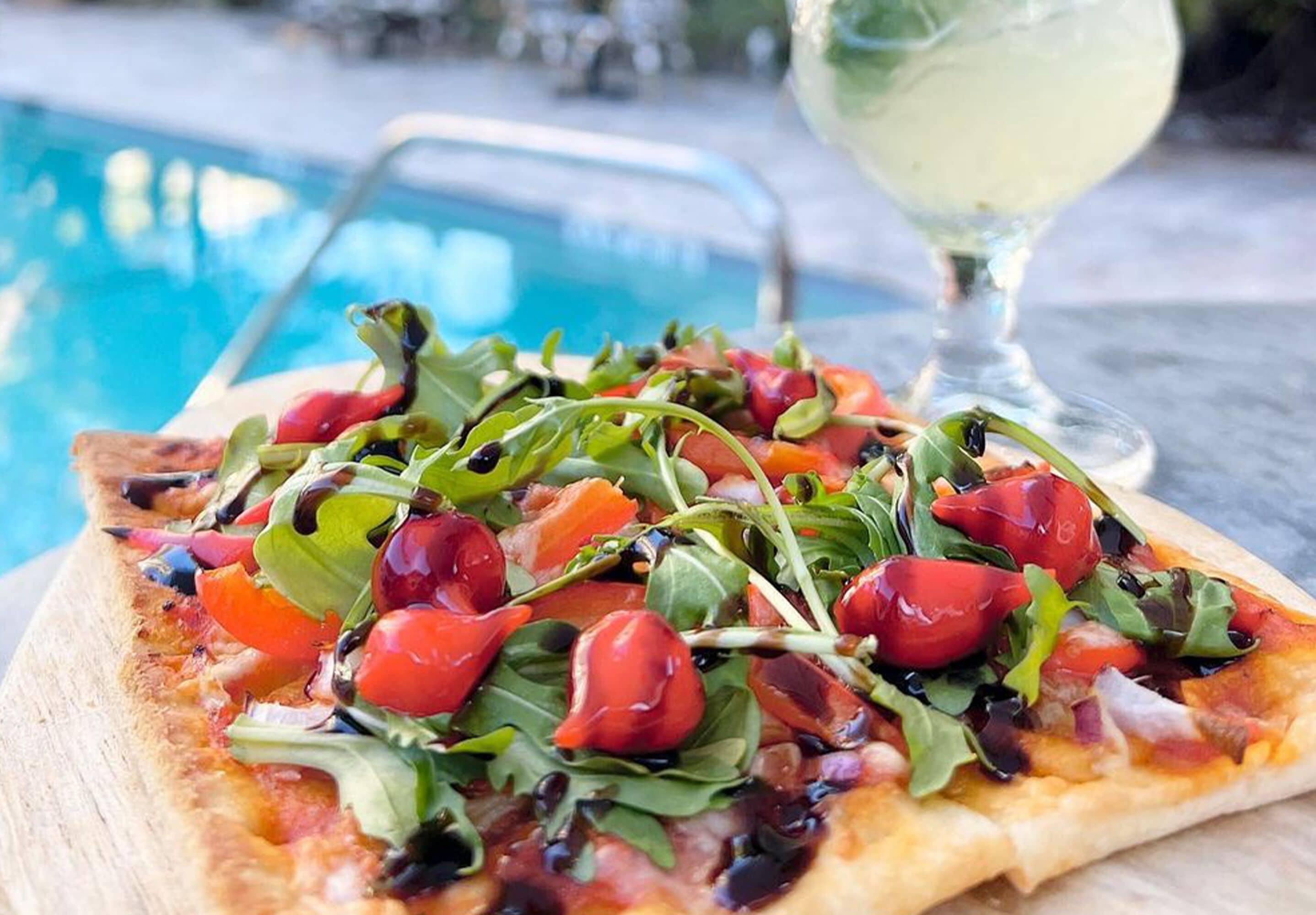 A pizza with fresh greens and tomatoes on a wooden board, a glass of white drink, and a pool in the background.