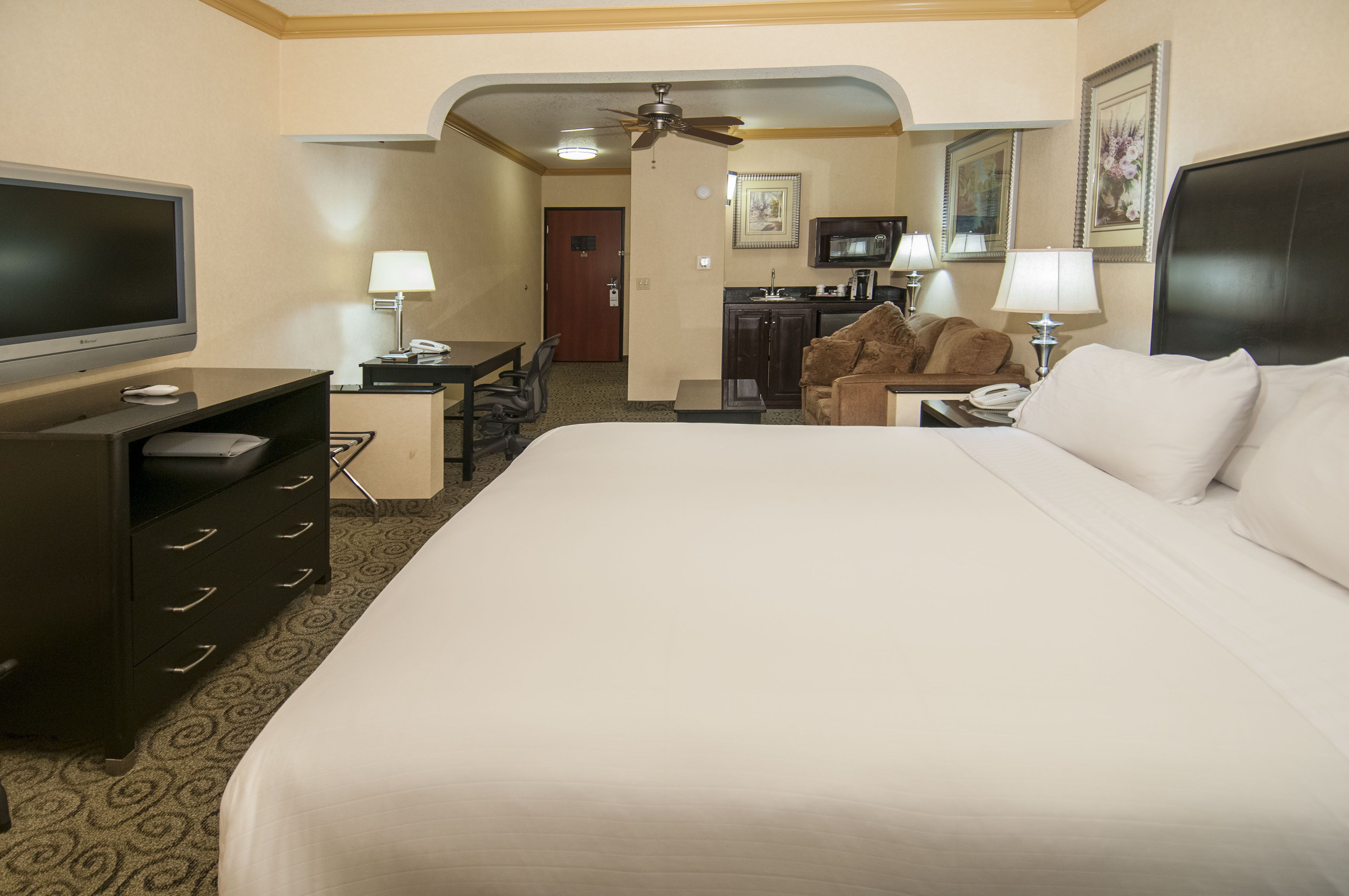 Quarto de hotel com uma cama grande, televisão, escrivaninha com cadeira, cozinha compacta e ventoinha no teto.