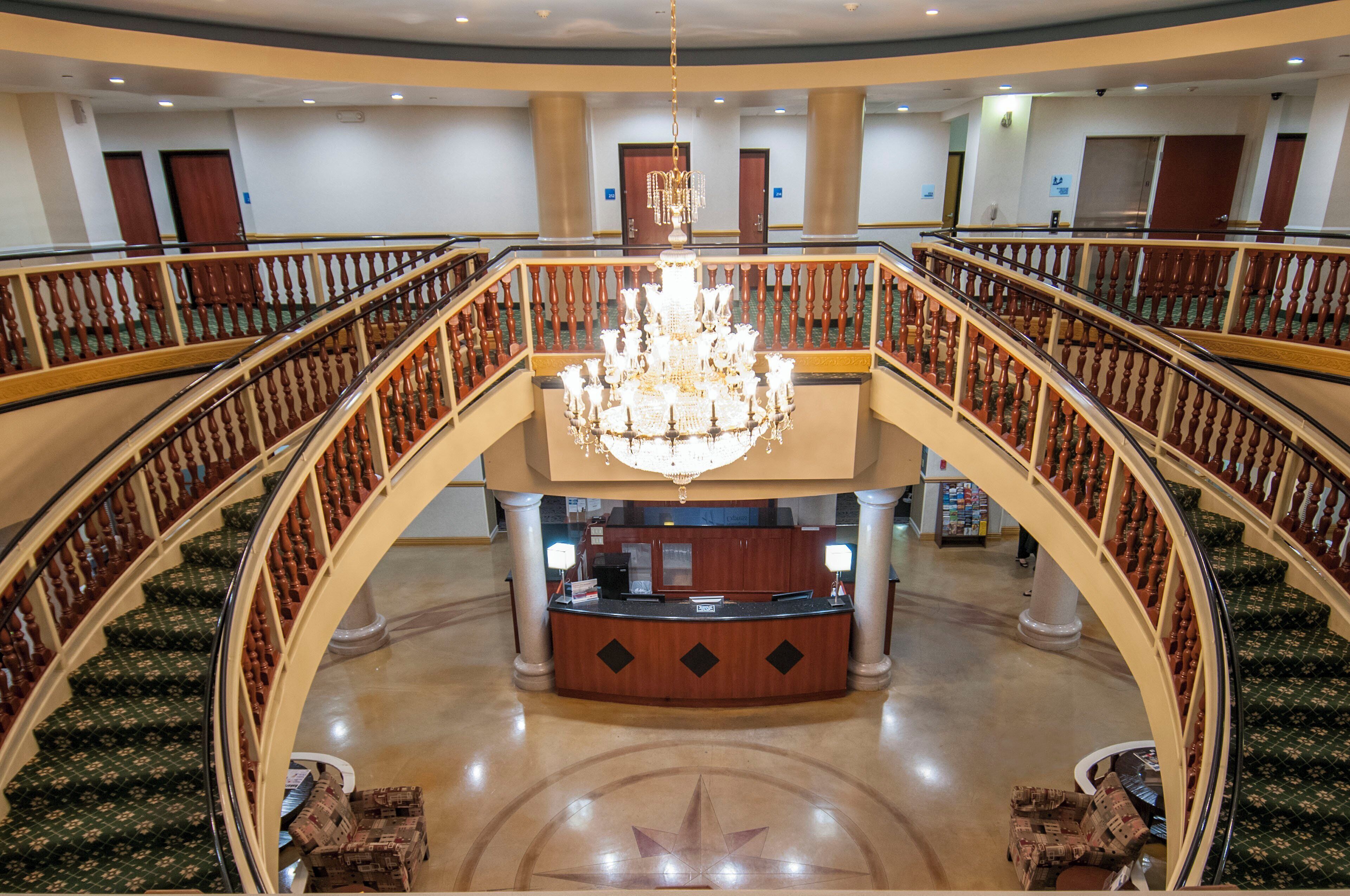 A grand lobby with a central chandelier, a reception desk, and a double staircase with wooden railings.