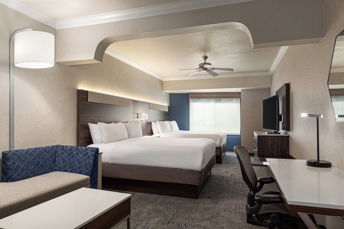 A hotel room with a large bed, a desk, a chair, a TV, and a ceiling fan.
