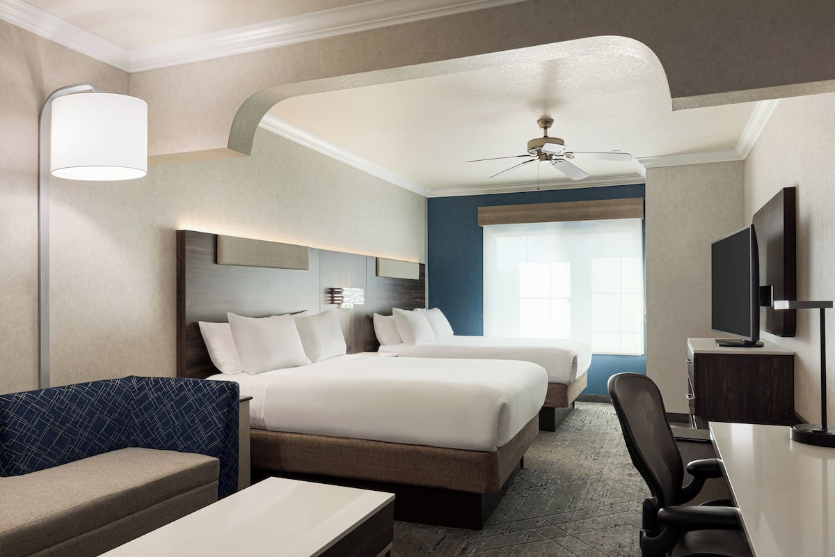 A hotel room with two beds, a desk, a chair, a TV, and a ceiling fan.
