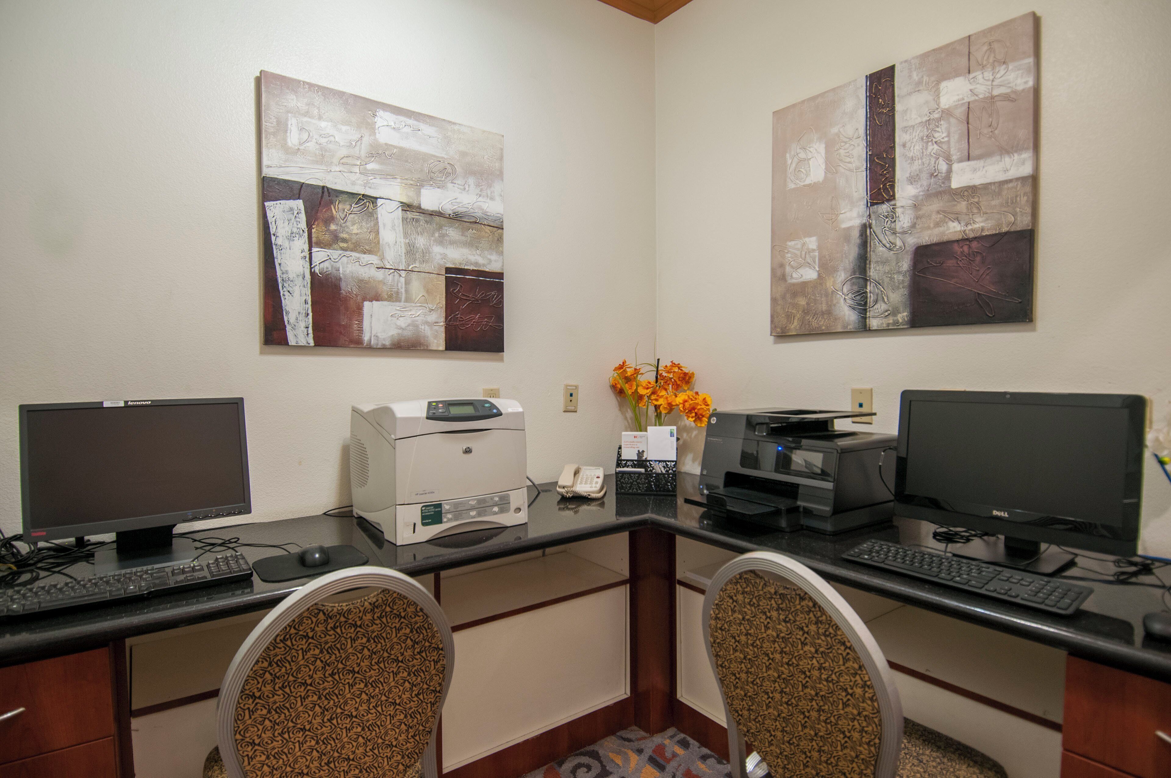A hotel office with two desks, each with a computer monitor, keyboard, and printer. There are two chairs in front of the desks, and a vase with flowers on the desk in the middle.