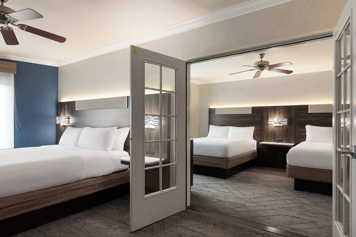 A hotel room with a large bed, a bedside table, a nightstand, a ceiling fan, and a door with a glass panel.