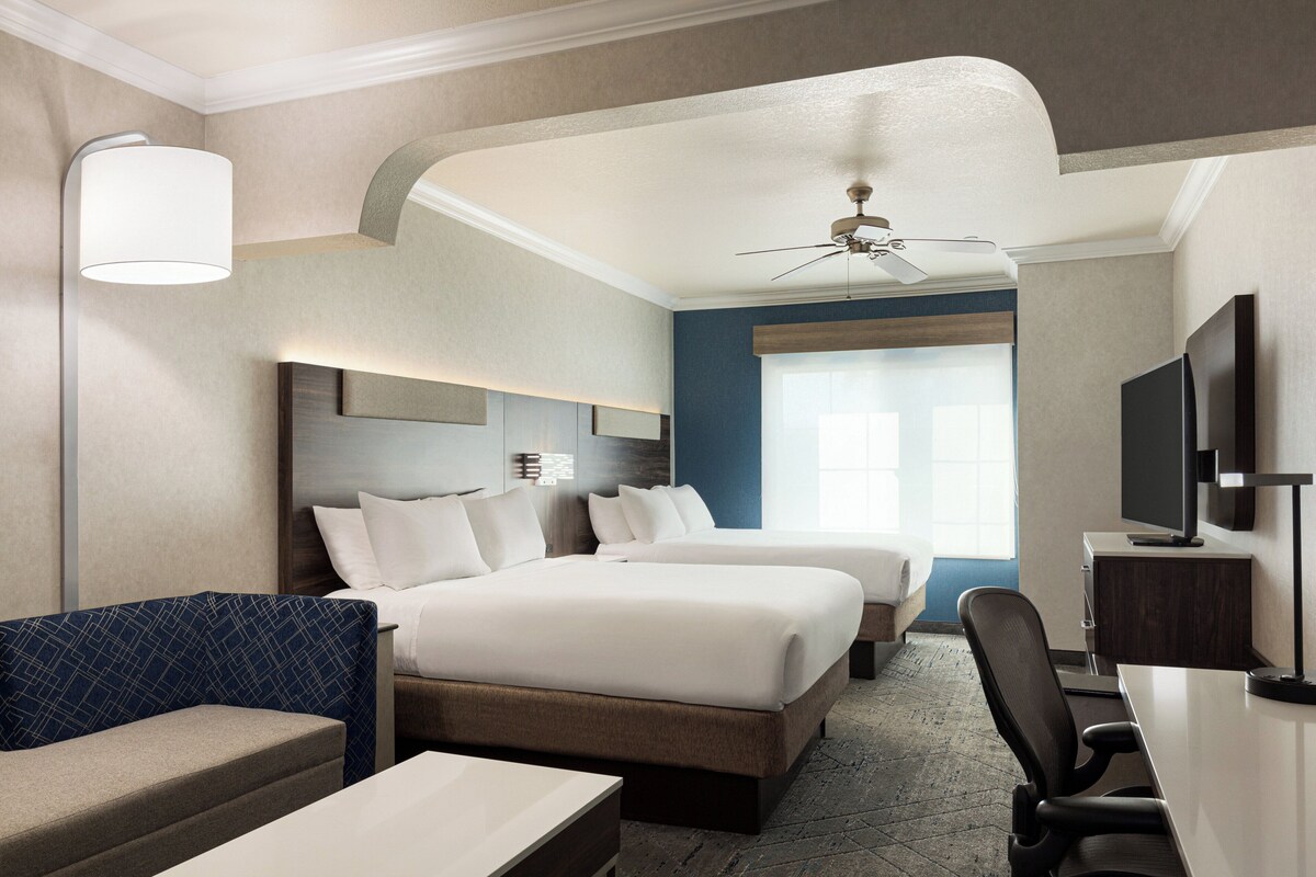A hotel room with two beds, a desk, a chair, a TV, and a ceiling fan.