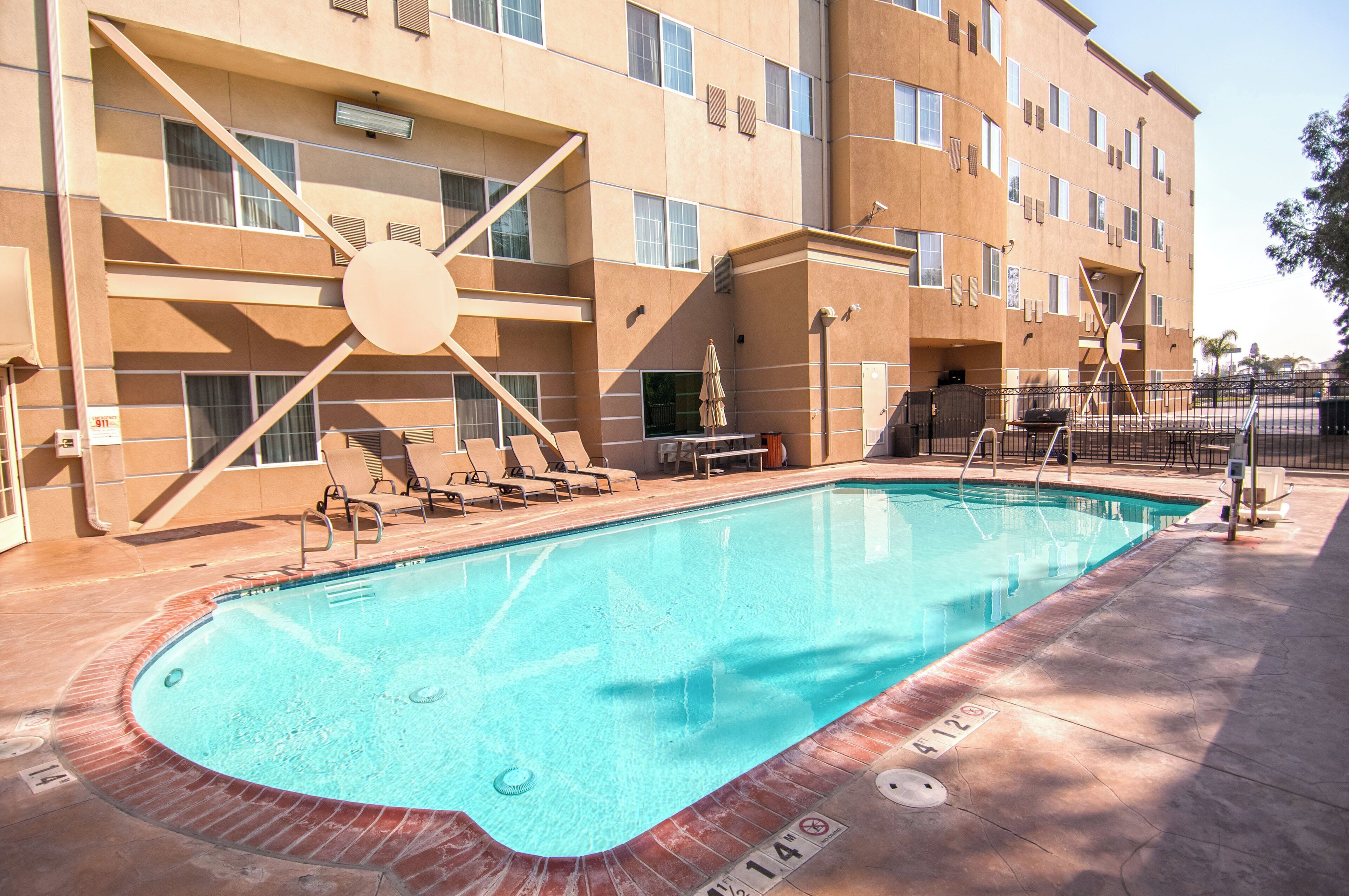 A swimming pool with lounge chairs and a sunshade, surrounded by a building with multiple windows and a fire alarm sign.