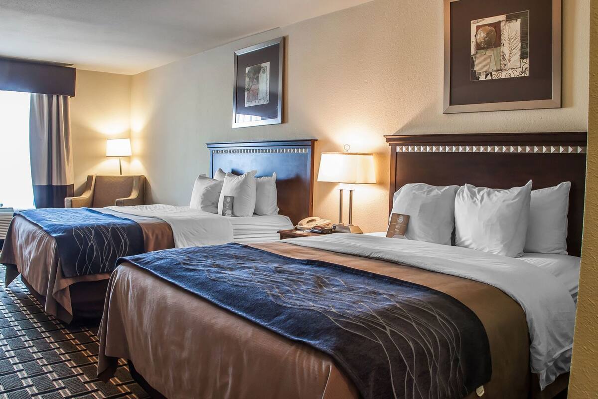 A hotel room with two beds, a desk, a chair, a lamp, and a framed picture on the wall.
