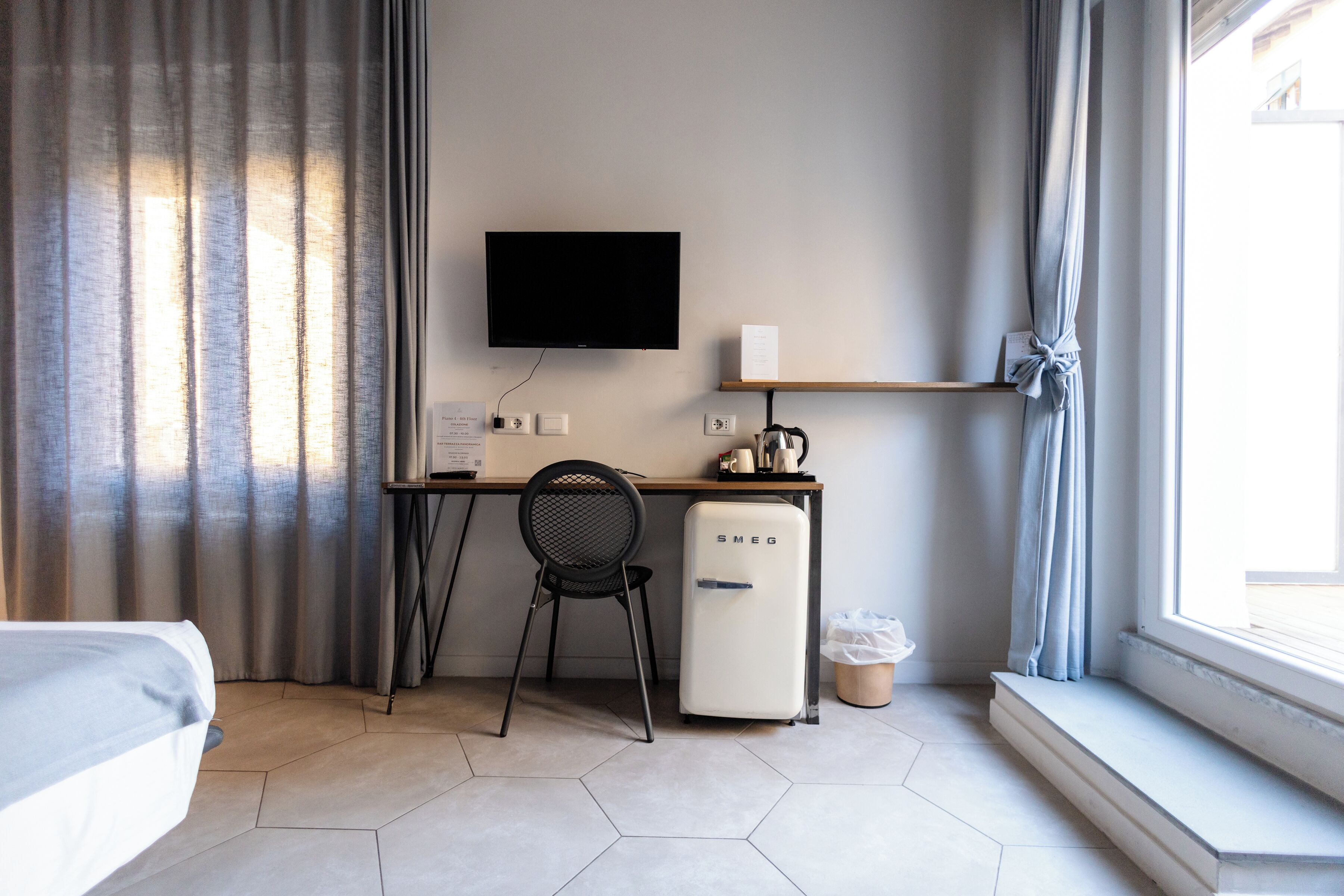 Habitació d'hotel amb un llit, una taula de treball amb cadira, televisió, nevera petita i una finestra amb cortines.