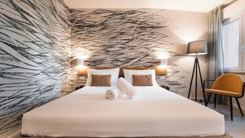 Una cama bien hecha con sábanas blancas y almohadas marrones, una lámpara de pie y una silla en una habitación con un estampado de hojas en la pared.