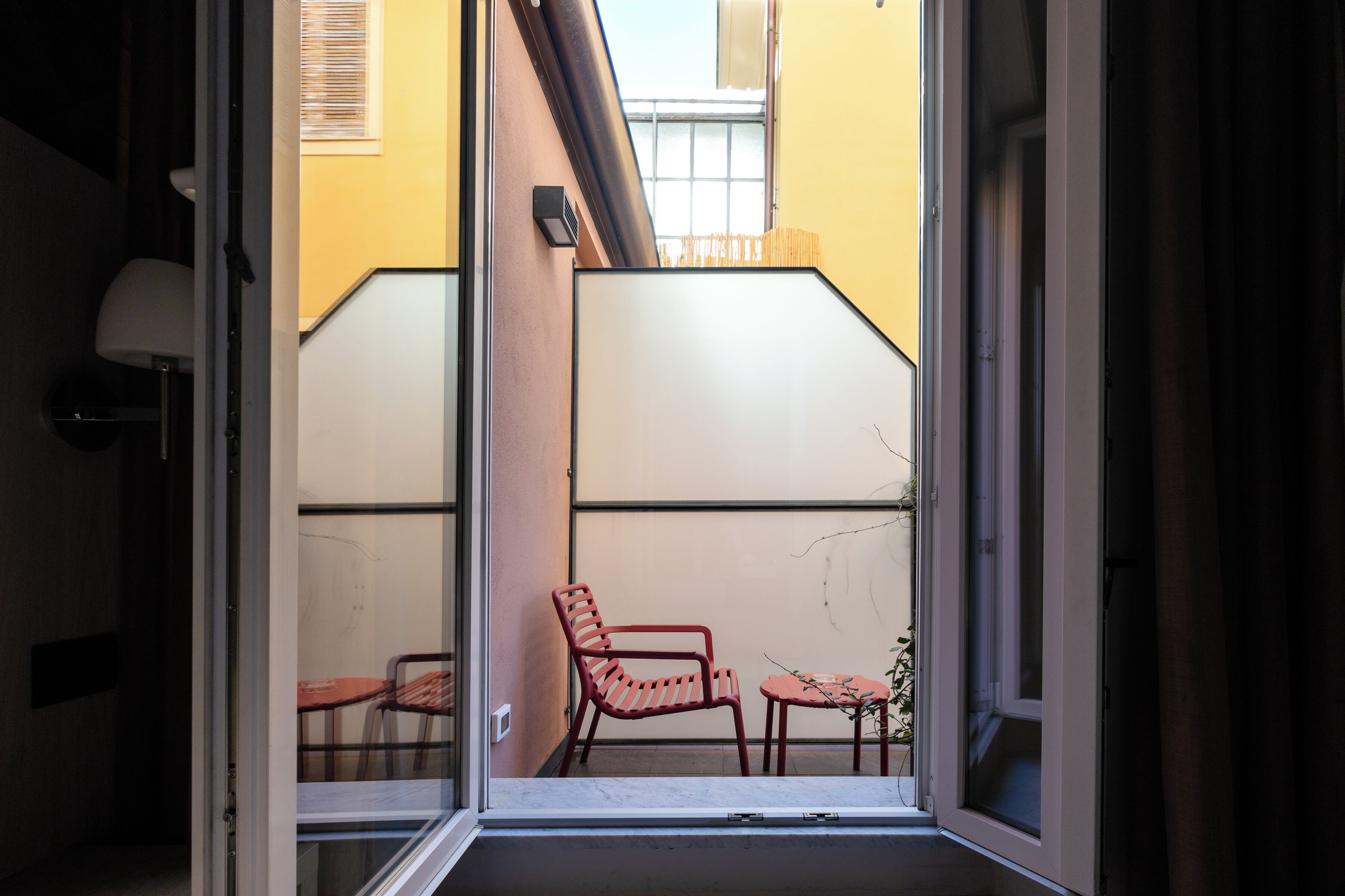 Ein Balkon mit einem roten Stuhl und einem kleinen Tisch, ein Fenster und Blick auf ein Gebäude mit einer gelben Wand und einer weißen Tür.
